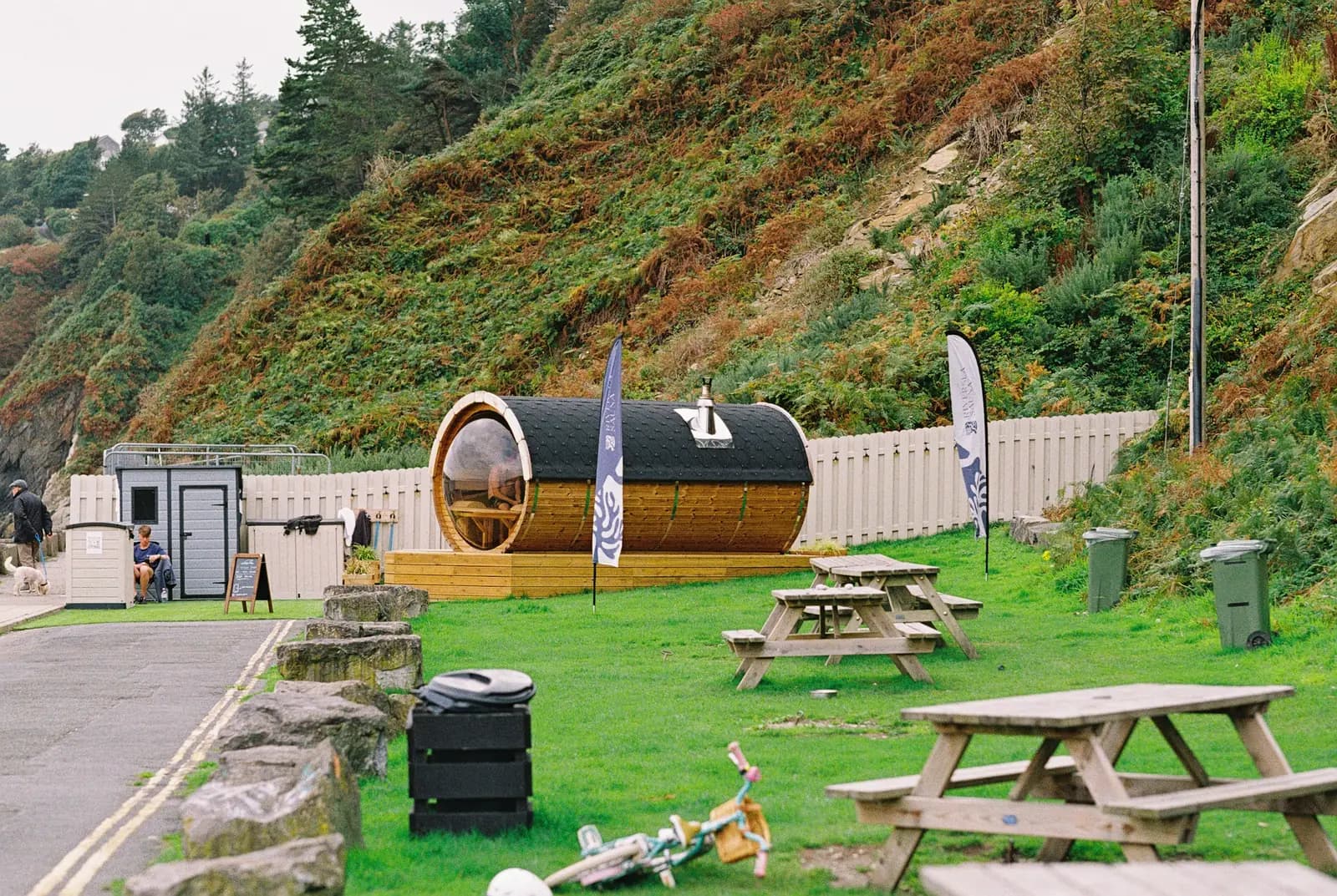 A barrel sauna on a grassy lawn below a steep green hillside, with picnic tables in front