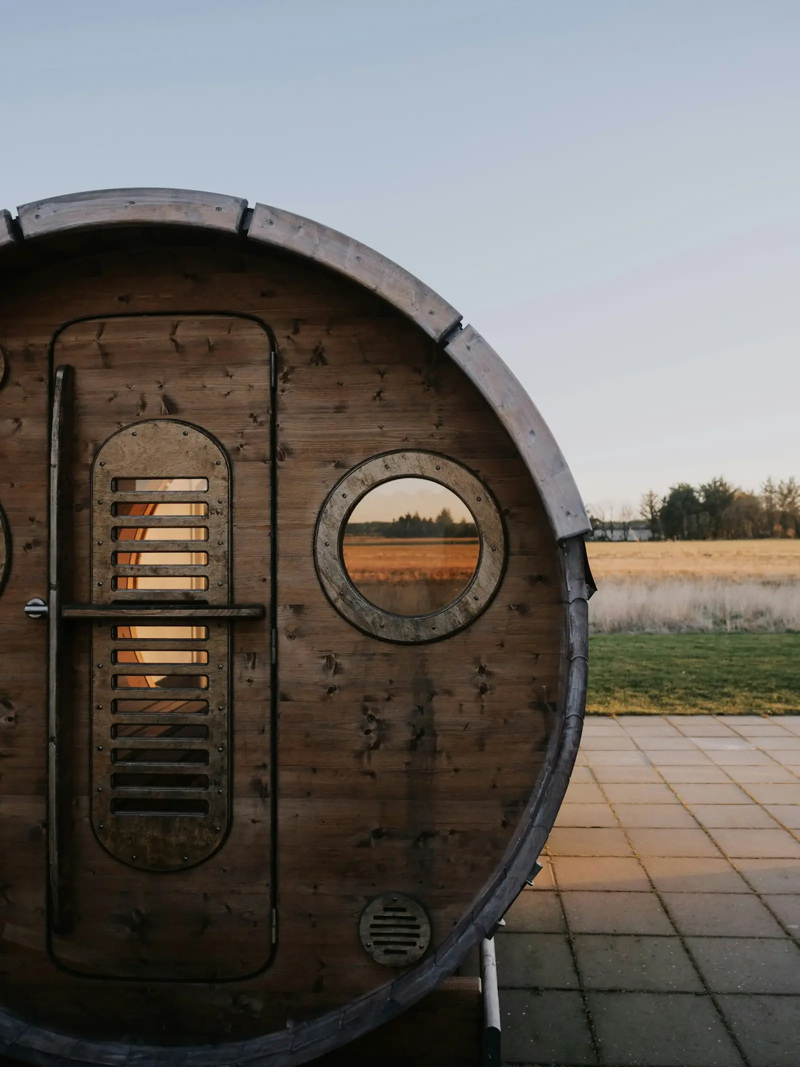The front of a barrel sauna with a round window, standing on a tiled patio
