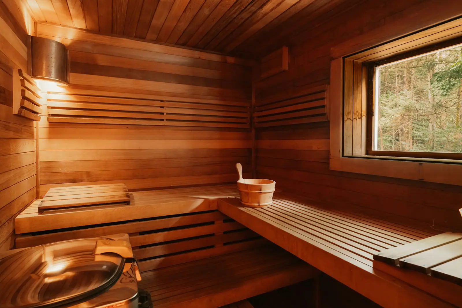 A wooden sauna with tiered benches, a bucket and a window onto trees