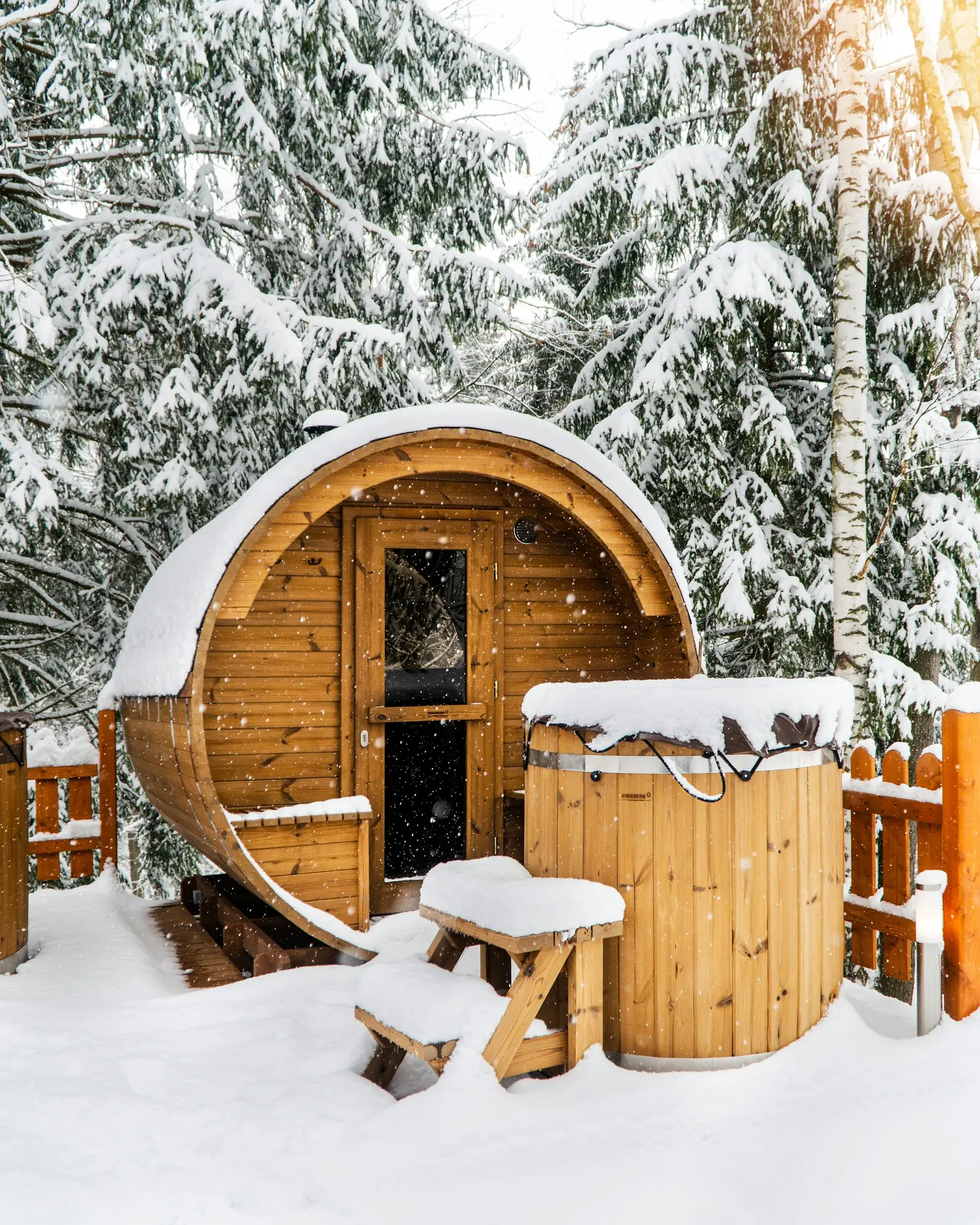 A barrel sauna and a wooden hot tub covered in snow among pine trees