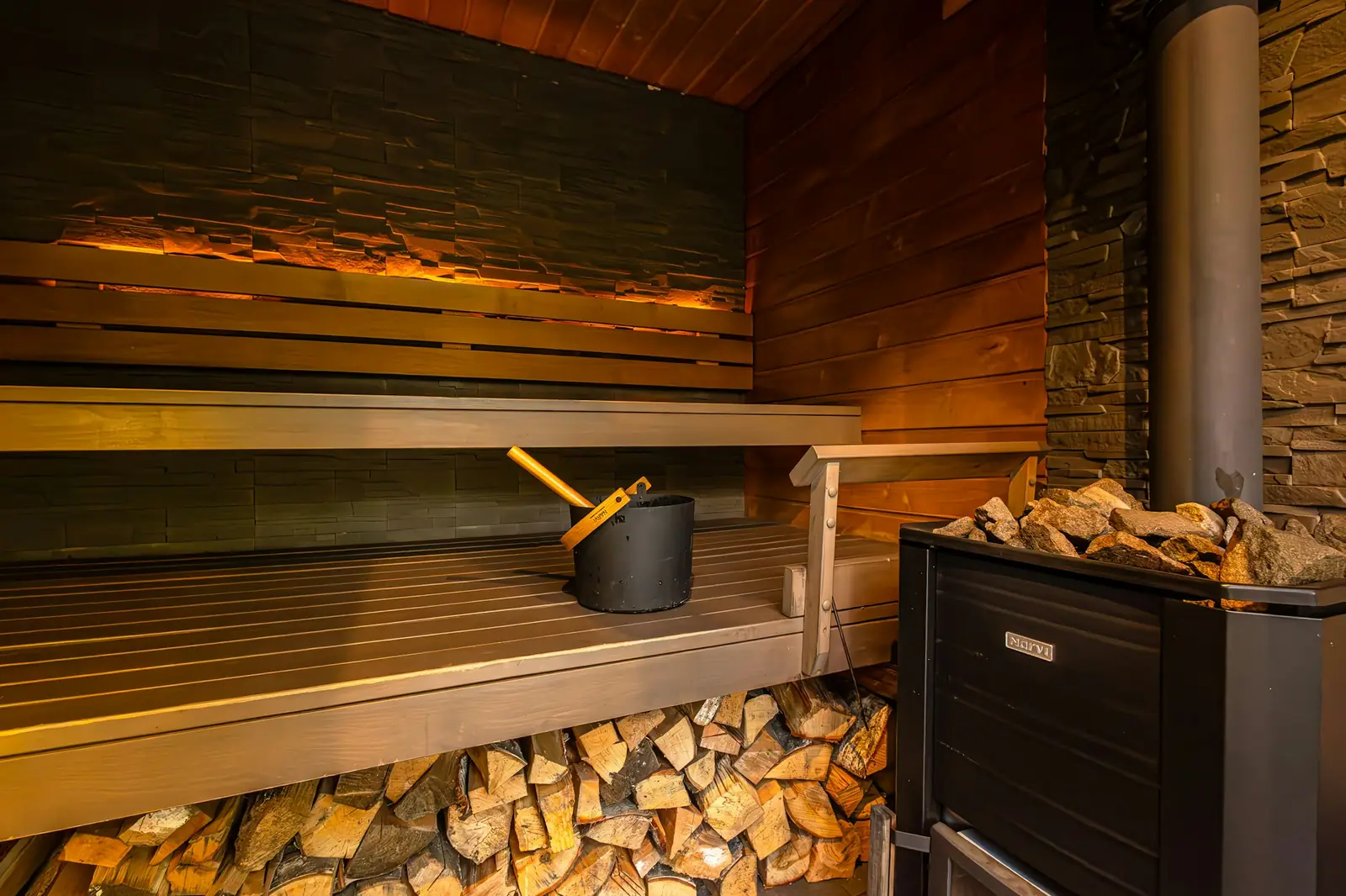 A sauna with a wood-burning heater, firewood stacked under the bench and a bucket on the bench