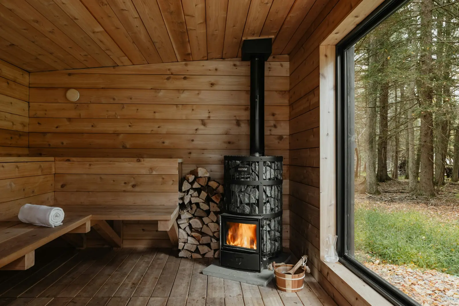 Inside a cabin sauna with flat walls, a wood-burning stove and a large window onto trees